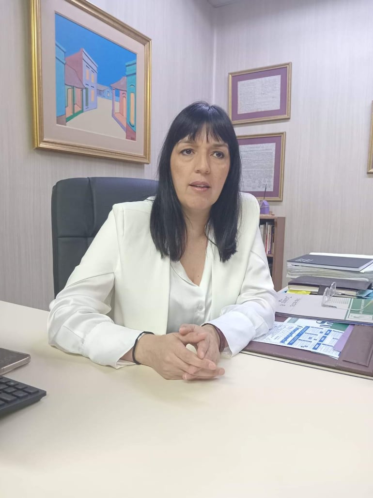 Mujer de cabello largo y oscuro en blusa blanca, sentada en un escritorio con documentos y teléfono móvil, en oficina luminosa.