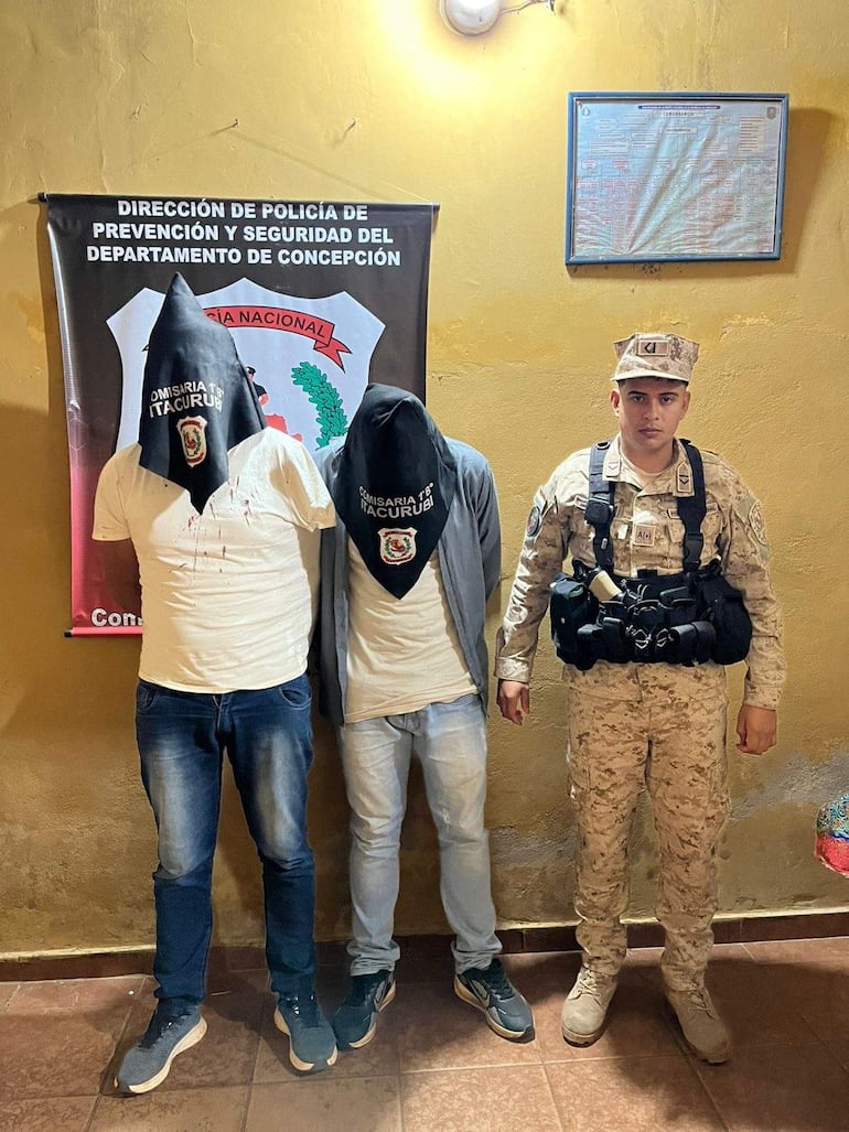 Dos aprehendidos encapuchados con camisetas blancas, custodiados por un policía con uniforme militar en un ambiente sobrio.