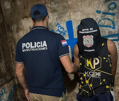 Agente policial en uniforme claro revisa a un individuo con capucha negra y maillot de Kobe Pharma en un ambiente descascarado y graffiteado.