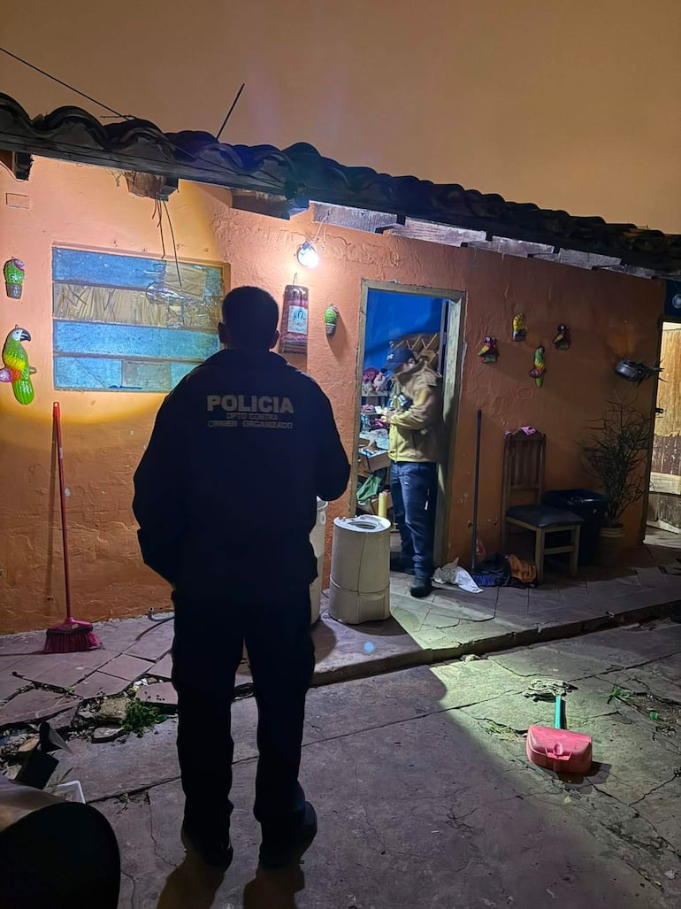 Oficial de policía de espaldas en chaqueta 'POLICÍA' mientras otra persona revisa objetos en una vivienda iluminada.