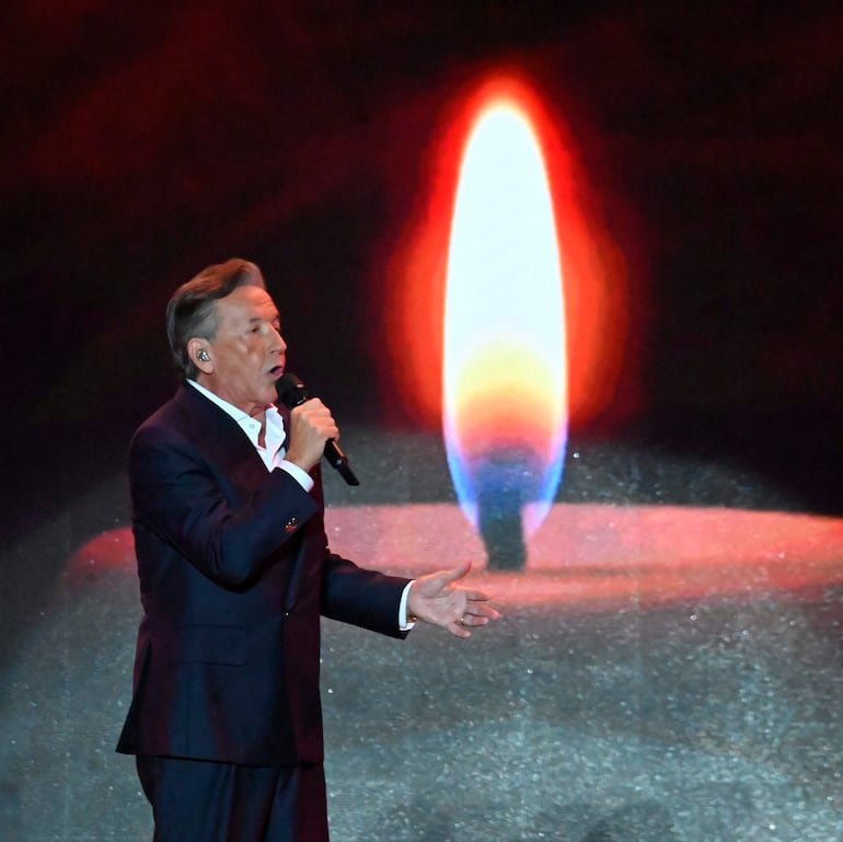 Ricardo Montaner canta con micrófono en mano, vistiendo traje oscuro y camisa blanca, bajo una iluminación cálida en el escenario.