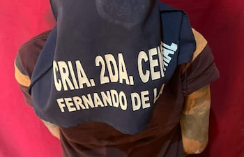 Hombre de espaldas con camiseta oscura y cabeza cubierta, en un entorno de color rojo, probablemente durante su detención.