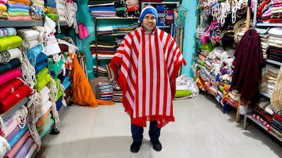 Hombre sonriente con poncho rojo y blanco en tienda repleta de mantas coloridas durante el festival.