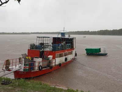 Barco rojo con la palabra 'MILA' en costado, rodeado de contenedores, en un ambiente lluvioso fluvial.