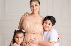 Nadia Portillo viste un vestido melocotón, sonríe mientras sostiene su abdomen; su hija, en vestido blanco, y su hijo, en camiseta blanca, sonríen también.