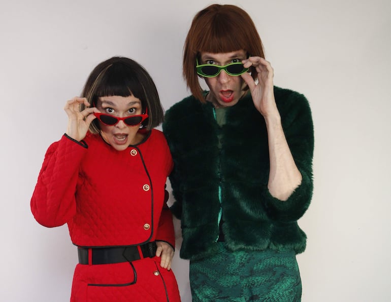 Lali González en abrigo rojo y Aldo Calabrese en pelaje verde, ambos con gafas de sol y expresiones sorprendidas en fondo neutro.