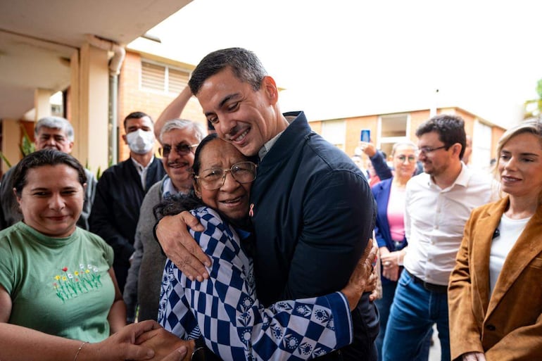 Santiago Peña en chaqueta oscura abraza a una mujer mayor con blusa de cuadros, ambos sonríen en un ambiente cálido.