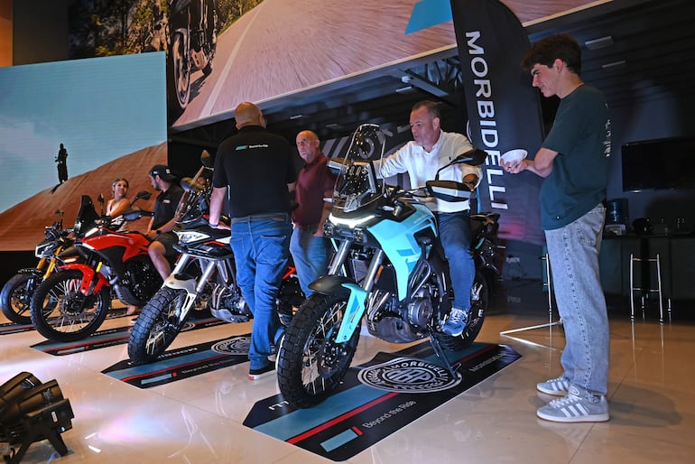 Hombre con motocicleta azul claro prueba el modelo, mientras otros conversan en un ambiente de presentación de Morbidelli.