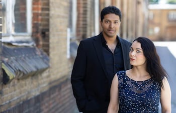Hombre en traje negro y mujer en vestido de noche azul posan en un entorno urbano con ladrillos de fondo.