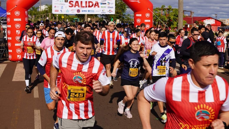 Participantes corren con camisetas a rayas rojas y blancas, con un ambiente festivo frente al arco de salida marcado como "SALIDA".