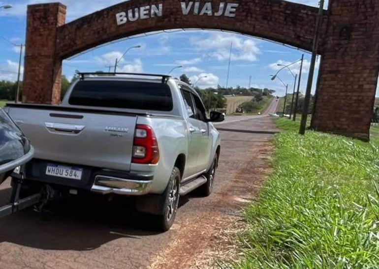 Camioneta plateada estacionada al lado de un camino, sin personas visibles. Arco 'BUEN VIAJE' de fondo y árboles alrededor.