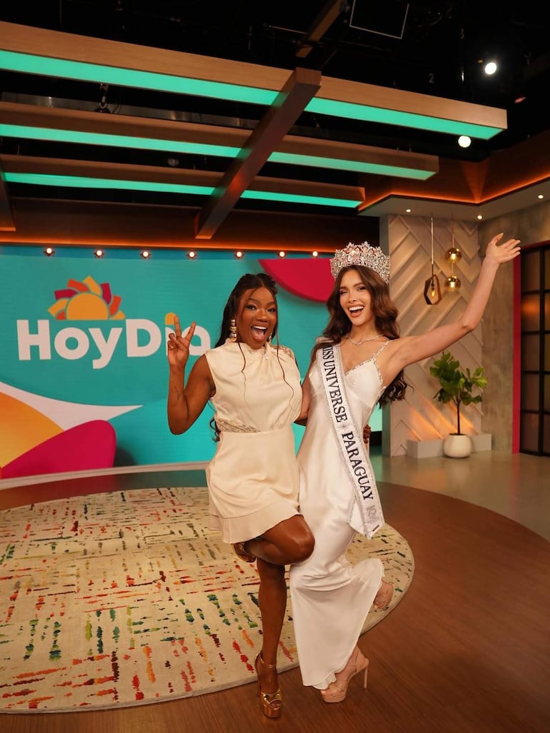 Gretha Matiauda sonríe con vestido claro, mientras la Miss Universe Paraguay luce vestido blanco y corona, ambas en un ambiente festivo.