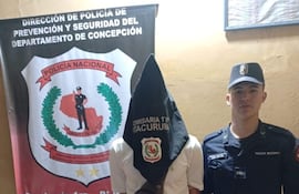 Hombre con bolsa en la cabeza, vestido con camiseta blanca y pantalones cortos, junto a un policía en uniforme negro en un ambiente interior.