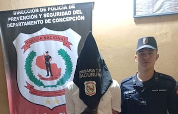 Hombre con bolsa en la cabeza, vestido con camiseta blanca y pantalones cortos, junto a un policía en uniforme negro en un ambiente interior.