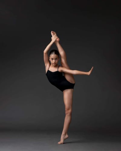 Bailarina en traje negro con pierna estirada, en un estudio iluminado suavemente, mostrando flexibilidad y control.