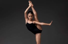 Bailarina en traje negro con pierna estirada, en un estudio iluminado suavemente, mostrando flexibilidad y control.