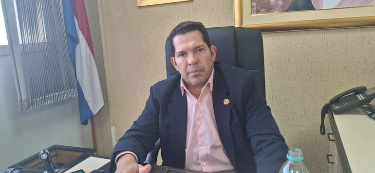 Hombre con saco negro y camisa clara sentado en oficina, con expresión seria y mirada hacia la cámara, rodeado de elementos de trabajo.