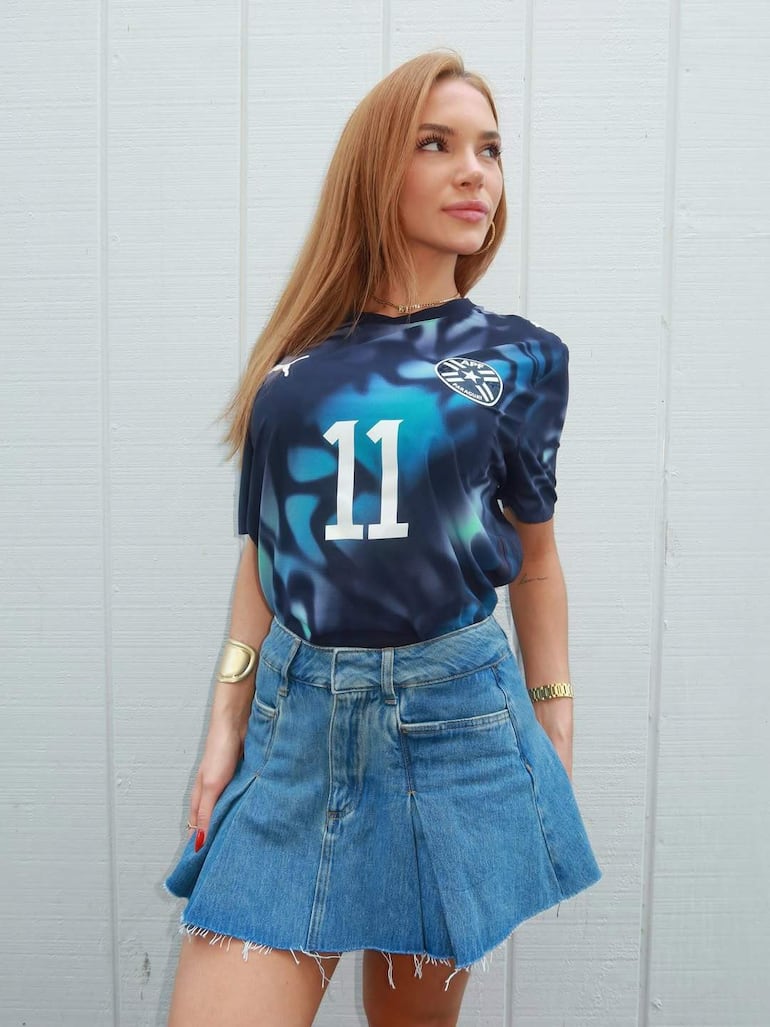 Mujer con cabello rubio, camiseta de fútbol azul con el número 11 y falda de mezclilla, posando de frente en fondo blanco.