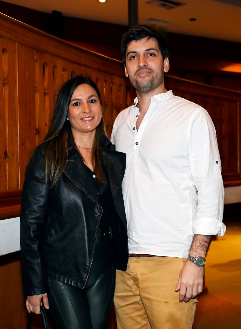 Mujer con chaqueta de cuero negra y pantalones oscuros sonríe junto a hombre con camisa blanca y pantalones beige en un ambiente elegante.