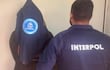 Agente de INTERPOL en chaqueta con emblema visible detiene a un hombre encapuchado en un ambiente formal.