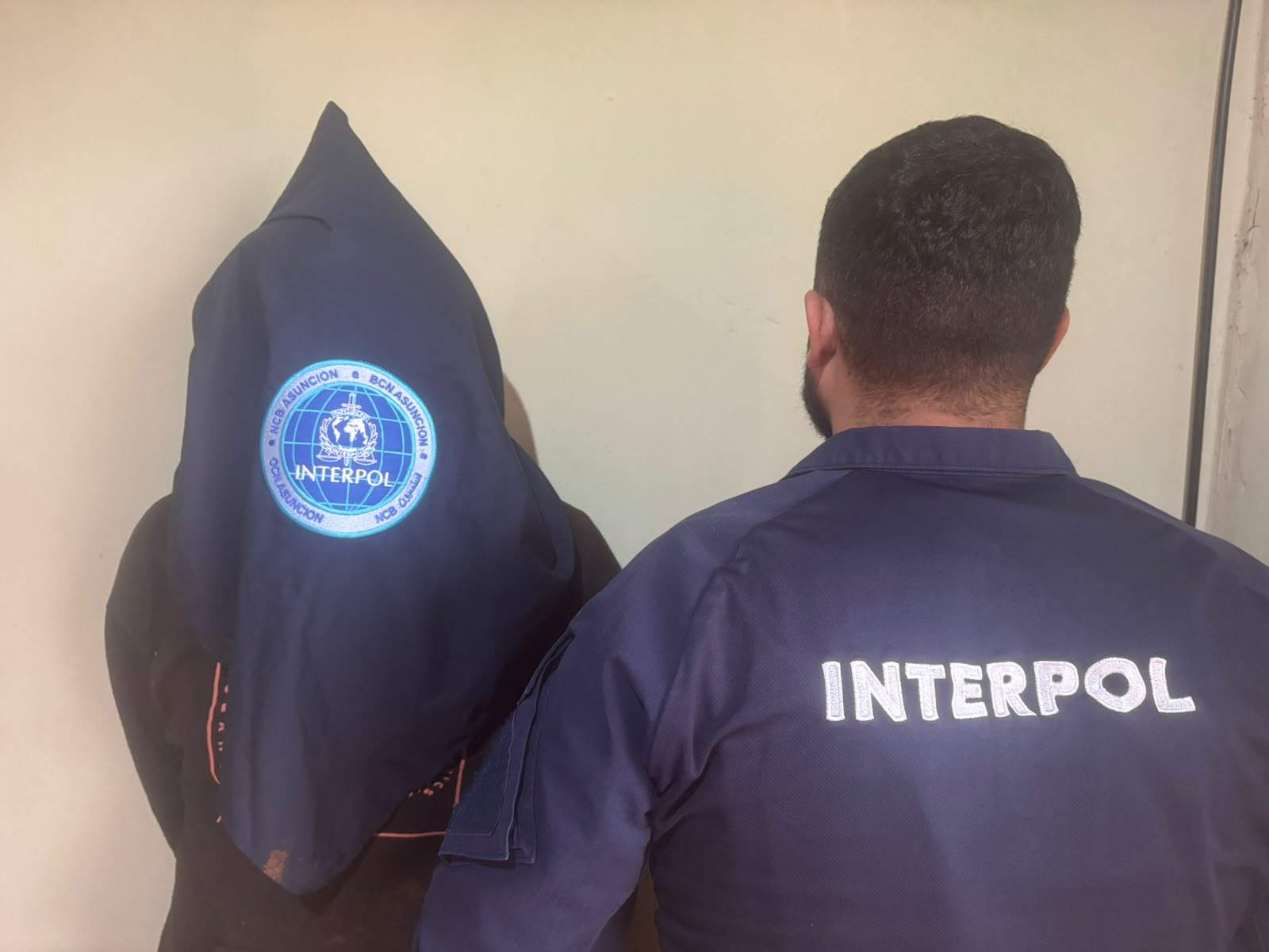 Agente de INTERPOL en chaqueta con emblema visible detiene a un hombre encapuchado en un ambiente formal.