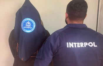 Agente de INTERPOL en chaqueta con emblema visible detiene a un hombre encapuchado en un ambiente formal.