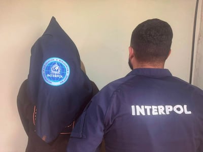 Agente de INTERPOL en chaqueta con emblema visible detiene a un hombre encapuchado en un ambiente formal.