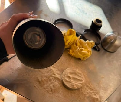 Mano sostiene un objeto cilíndrico en una superficie de cocina con polvo y utensilios dispersos.