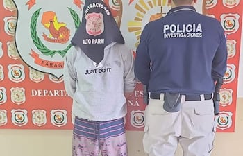 Individuo de espaldas con capucha negra y camiseta gris, junto a un policía en uniforme en un ambiente interior.