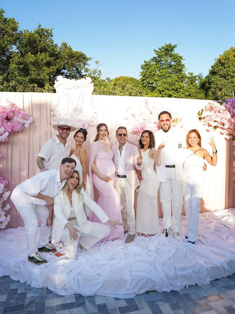 Nadia Ferreira y Marc Anthony sonríen rodeados de ocho personas en un ambiente festivo, todos con ropa clara y decoración floral de fondo.