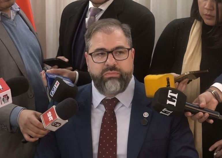 Rodrigo Nicora con traje azul y corbata de puntos, rodeado de periodistas con micrófonos en un ambiente formal.