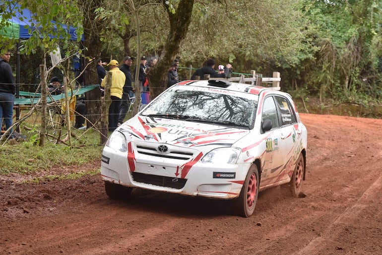 Espectadores con chaquetas y sombreros observan un Toyota de rally rojo avanzando en circuito de tierra y vegetación.