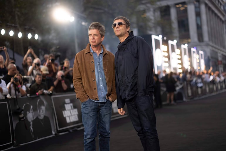 Noel y Liam Gallagher en chaquetas, posando con tranquilidad en una alfombra roja rodeados de fotógrafos.