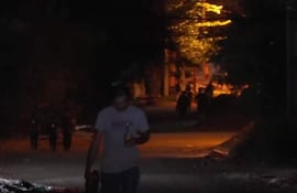 Hombre con camiseta gris y pantalones oscuros camina sosteniendo un objeto, en una calle poco iluminada con árboles y edificaciones.