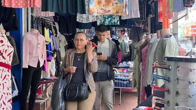 Mujer de mediana edad con gafas y blusa oscura usa su móvil, mientras un joven en camiseta clara está enfocado en su teléfono.