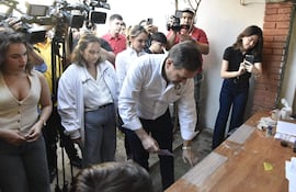 Hombre con camisa blanca trabajando en mesa, mujeres observando atentamente. Ambiente informal con varios asistentes de camisa oscura.