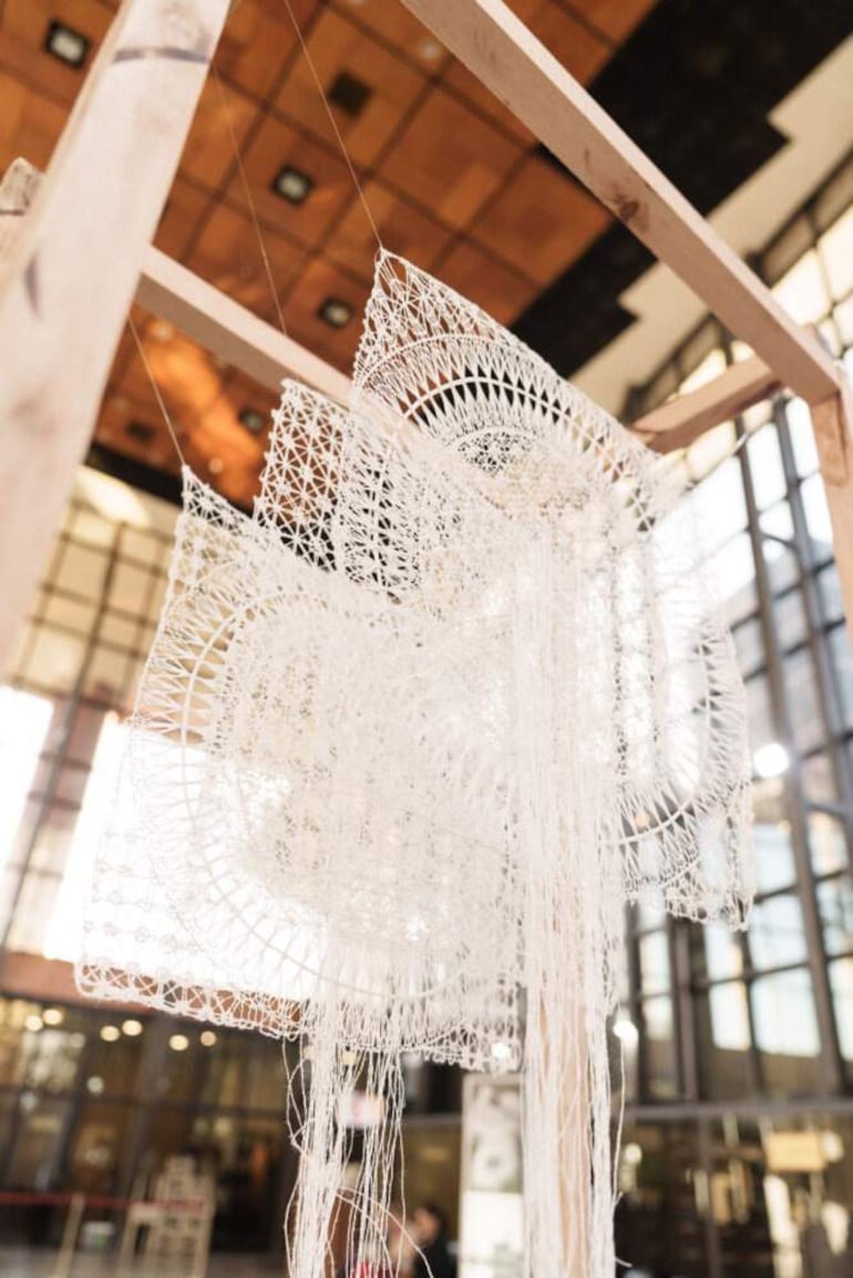 Estructura de exhibición con obras de encaje blanco colgando de un marco de madera en un espacio cultural iluminado.