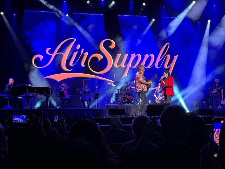 Air Supply en el escenario, dos cantantes con camisa colorida y traje rojo, rodeados de músicos y luces brillantes durante un emocionante concierto.