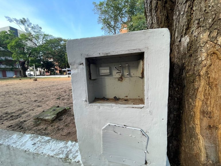 Caja de conexión eléctrica blanca, ubicada junto a un árbol en un entorno urbano con edificios al fondo.