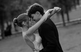 Mujer en vestido blanco y hombre en traje oscuro abrazándose en un parque, sonriendo y mirando el uno al otro.