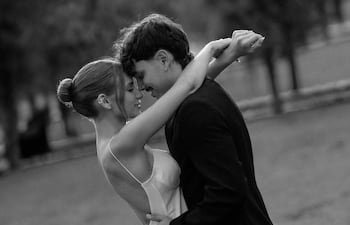 Mujer en vestido blanco y hombre en traje oscuro abrazándose en un parque, sonriendo y mirando el uno al otro.