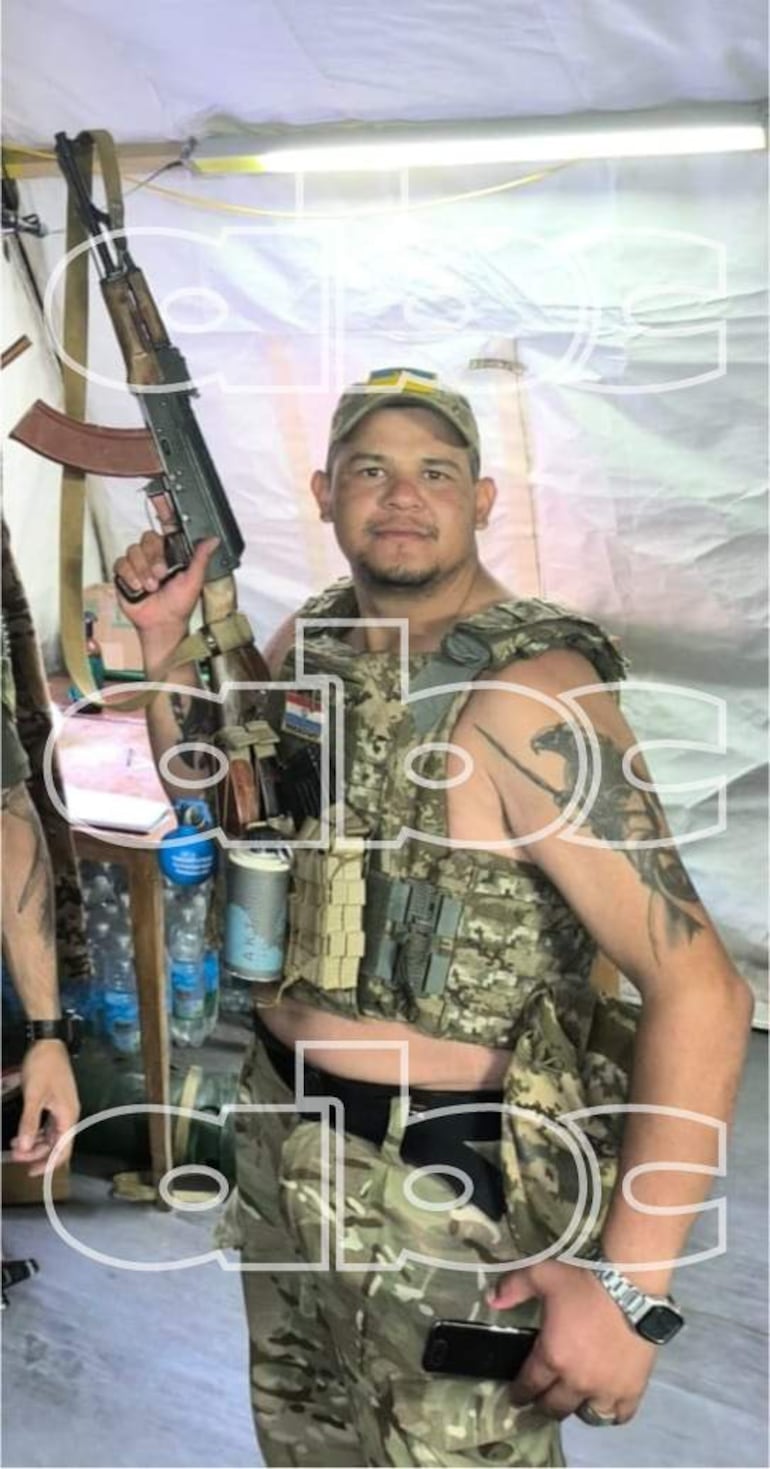 Eduardo Román en uniforme militar con un arma, exhibiendo una expresión tranquila en un entorno militar.
