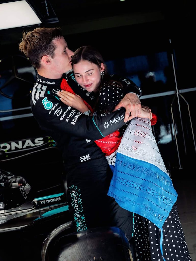 Joshua Duerksen en traje de piloto abrazando a Anneke Rahn, vestida con un vestido de lunares, en un taller con un coche de carreras de fondo.
