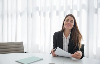 Mujer profesional con traje ejecutivo sonriendo en una oficina, sentada frente a un escritorio con documentos, representando la selección de personal y el éxito laboral en ABC Empleos.