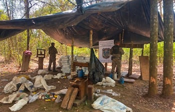 Dos agentes militares en vestimenta táctica operan bajo una lona negra en un campamento, rodeados de sacos y latas.