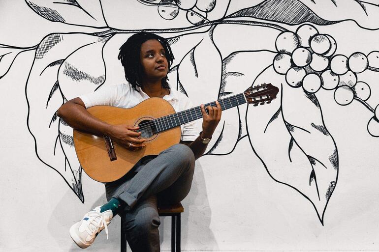 Gabriele Leite con cabello rizado, camisa blanca y pantalones grises, tocando guitarra en un café decorado con un mural.