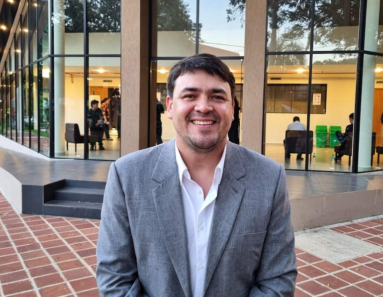 Benítez Herebia, sonriente, viste saco gris y camisa blanca, con un entorno de edificios modernos y personas en interior.