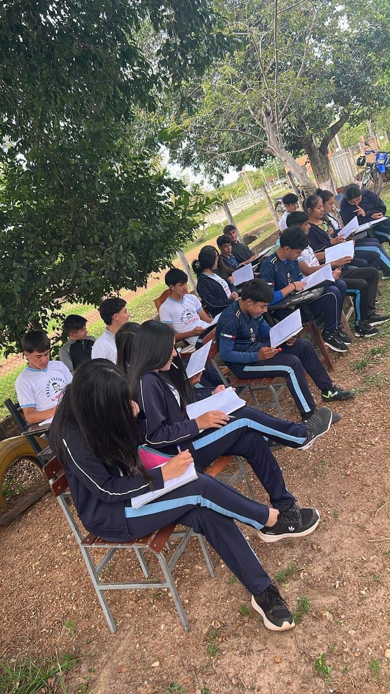 Grupo de estudiantes en uniformes deportivos oscuros con detalles en azul, tomando notas bajo un árbol en un ambiente agradable.