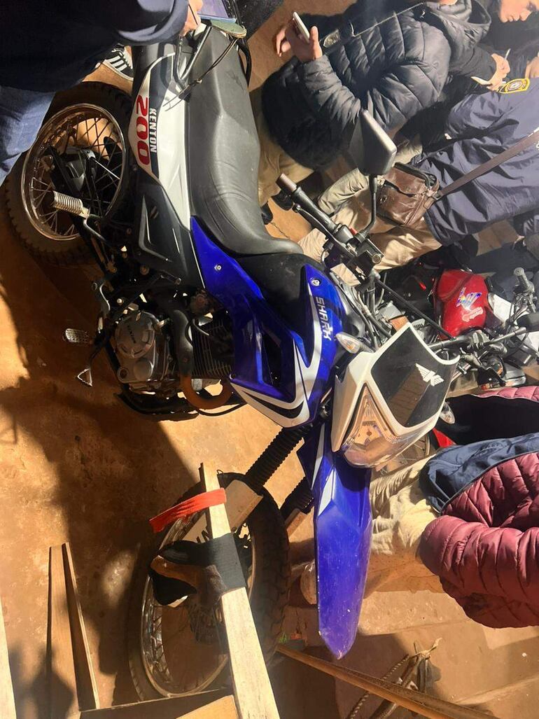 Grupo de personas en un garaje, examinando una motocicleta Kenton azul y usando teléfonos móviles.