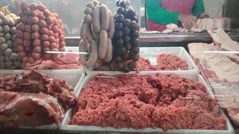 Puesto de venta con bandejas de carne picada, trozos grandes y chorizos colgados, en ambiente de carnicería.
